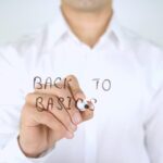 A man in a white shirt writing the words Back to Basics with a marker on a transparent glass board.