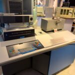 An IBM 1130 computing system on display in a museum, with its console panel, typewriter and keyboard on a grey and blue desk.