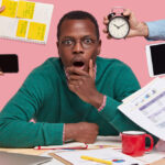 A startled man in glasses and a green jumper sitting at a desk while hands around him push a phone, a tablet, an alarm clock, a notebook and documents at him.