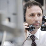 A businessman in a white shirt and tie holding a phone to each ear, listening to two calls at once.
