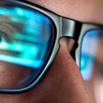 A close-up of a person's eye behind glasses, with lines of code from a screen reflected in the lenses.
