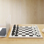 A chessboard on a wooden desk with a laptop playing the black pieces and a model of a human brain playing the white pieces.