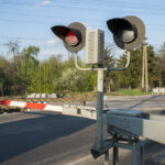 A closed railway level crossing, with its red and white barrier lowered across the road and a red signal light on.